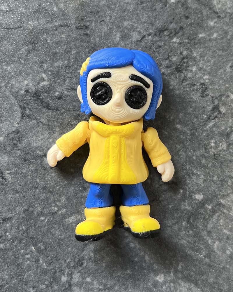 Coraline mini figures with eerie black button eyes, yellow raincoat, and blue hair, inspired by gothic stop-motion style.